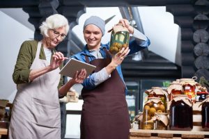 Cheerful handsome young grandson in apron helping concentrated grandmother with stock-taking in shop, they examining preserves using tablet